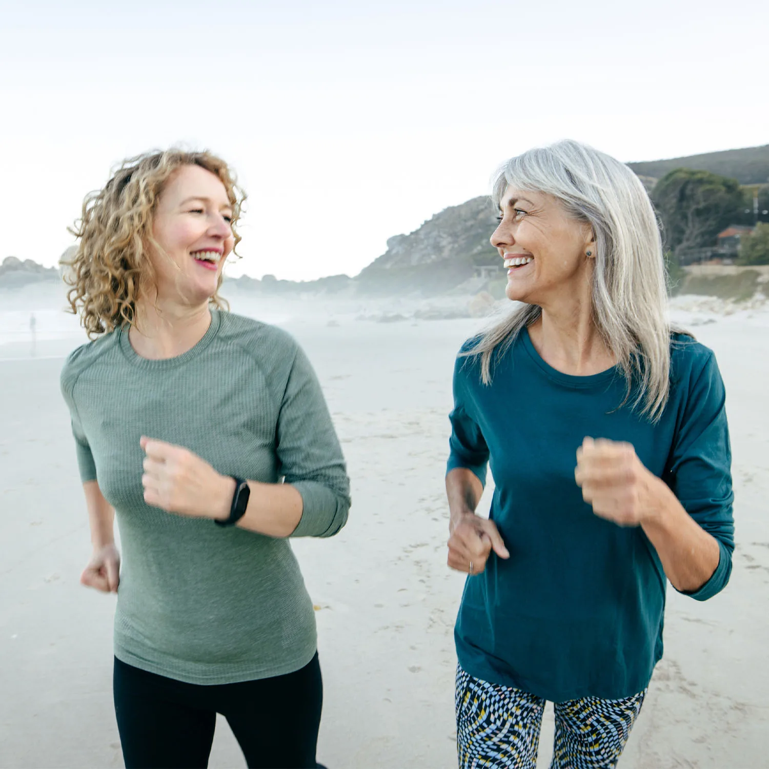 Zwei ältere Damen joggen am Strand entlang, lachen sich dabei gegenseitig an und strahlen.