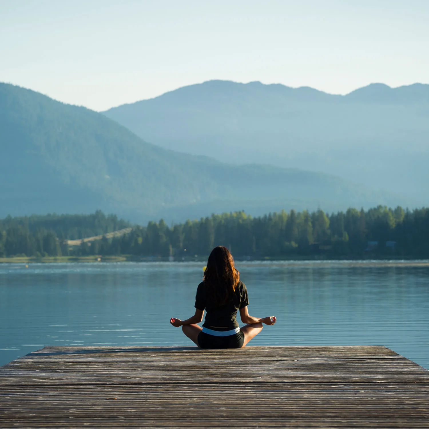 Eine Frau von hinten, die meditierend auf einem Steg an einem See sitzt.