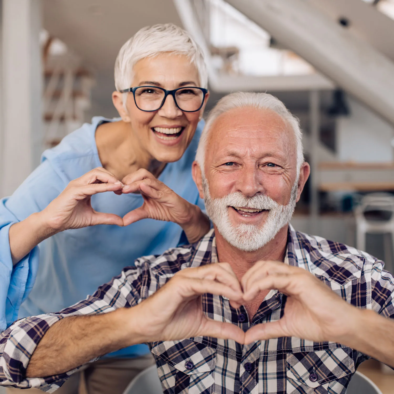 Eine ältere Frau und ein älterer Mann, die zusammen lachen und ein Herz mit ihren Händen formen.
